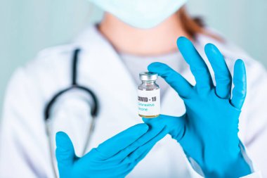 Üniformalı kadın doktor veya hemşire ve yüz maskesi takan eldiven laboratuarda koruyucu ilaç şişesinde COVID-19 Coronovirus aşı etiketi taşıyor.