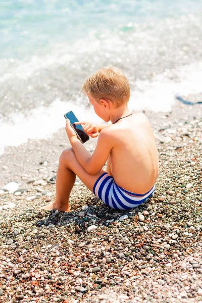 小さな男の子はビーチで電話でゲームをしている 海辺の概念で休暇中の子供のためのガジェット依存障害の問題 — ストック写真