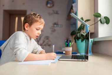 Evde ödevlerini yapan sevimli bir okul çocuğu. Eğitim kitapları ve defter masanın üstünde. Uzaklık öğrenimi çevrimiçi eğitim