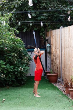 Güneşli bir arka bahçede duran genç bir çocuk ok ve yayla okçuluk çalışıyor. Yeşil çimenler ve çiçeklerle çevrili olan o, hedefine dikkatle odaklanır. Sahne, konsantrasyon, yetenek geliştirme ve dışarıda oynamanın huzurlu bir şekilde eğlencesini yakalar.