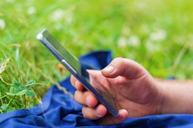 Şehir parkında yeşil çimlerin üzerinde uzanan akıllı telefonu tutan yetişkin bir adam.
