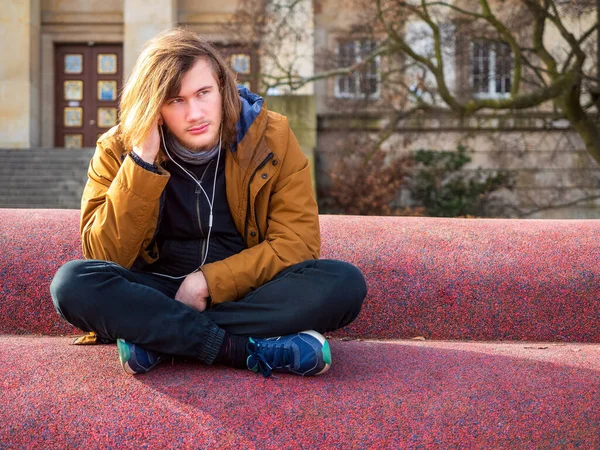 Üzgün genç bir adam şehirde tek başına oturuyor ve müzik dinliyor.