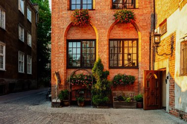 Sıcak, eski Avrupa şehri dar sokağı ve güneşli bir günde tuğla bir duvarı kaplayan çiçekli eski binanın cephe manzarası. Gdansk, Polonya