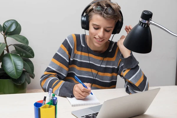 Smiling teenager in glasses and headphones with pen in hand studying online with teacher on laptop