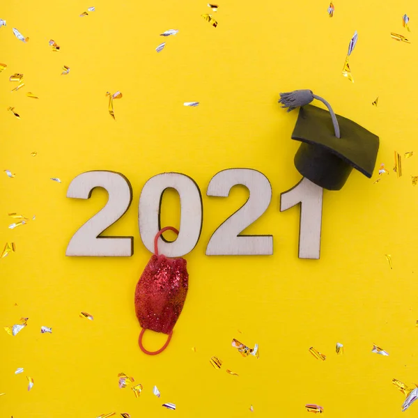 Graduation during coronavirus 2021: wooden number 2021 in graduation hat with medical mask and confetti on yellow background.