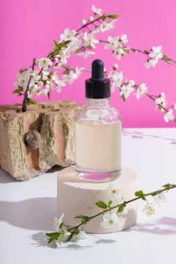 A glass dropper bottle on a podium with blooming twigs on a pink background. Vertical stock photo