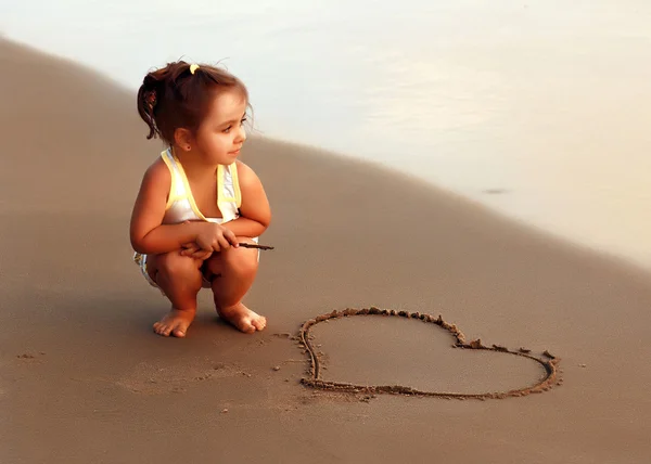 Pensive girl sitting at the evening beach near painted heart and looks into the distance at sea — Stock Photo, Image