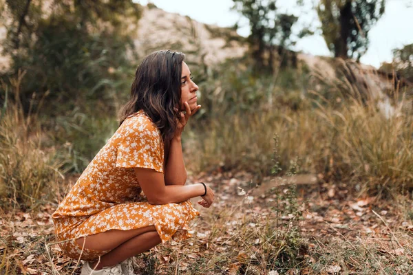 Nachdenkliche schöne brünette Frau in einem Kleid hockt im Wald — Stockfoto