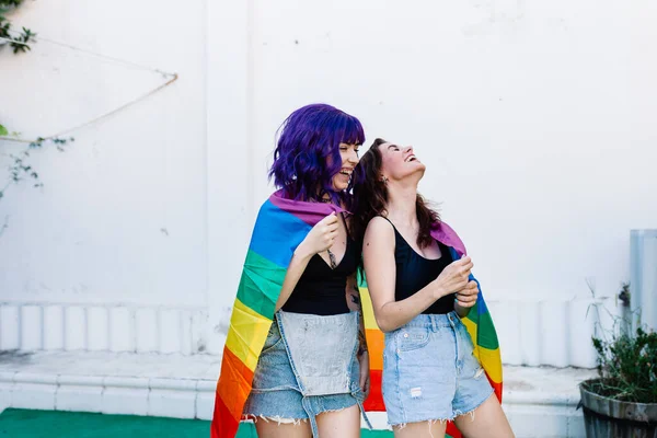 Dos mujeres con bandera de arco iris en un patio trasero. Pareja Chicas lesbianas jóvenes sosteniendo una bandera de arco iris y abrazándose felizmente. Un par de mujeres lesbianas — Foto de Stock