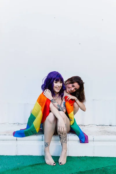 Dos mujeres con bandera de arco iris en un patio trasero. Pareja Chicas lesbianas jóvenes sosteniendo una bandera de arco iris y abrazándose felizmente. Un par de mujeres lesbianas — Foto de Stock