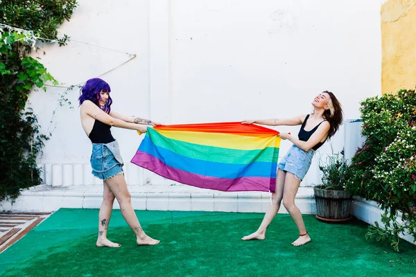 Dos mujeres con bandera de arco iris en un patio trasero. Pareja Chicas lesbianas jóvenes sosteniendo una bandera de arco iris y abrazándose felizmente. Un par de mujeres lesbianas — Foto de Stock