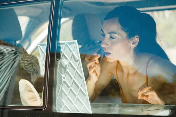 Donna attraente in bikini, si guarda allo specchio mentre fa il trucco all'interno del suo camper, concetto di viaggio su strada — Foto Stock