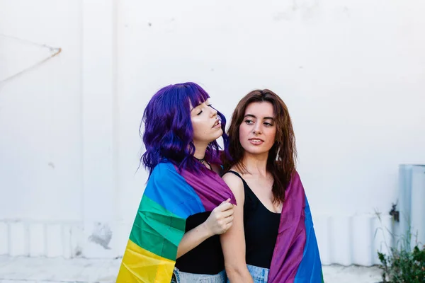 Dos mujeres con bandera de arco iris en un patio trasero. Pareja Chicas lesbianas jóvenes sosteniendo una bandera de arco iris y abrazándose felizmente. Un par de mujeres lesbianas — Foto de Stock