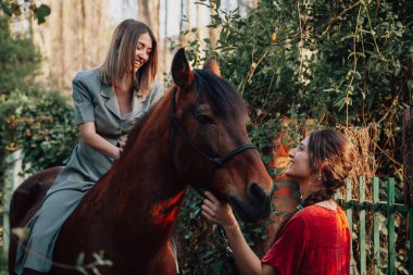 İki kadın arkadaş sohbet ediyor ve atlarıyla kırsalda geziyorlar.