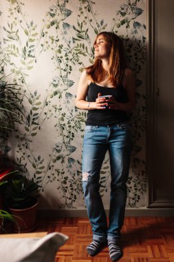 Genç kızıl saçlı kız, elinde fincanla pencerede duruyor ve evde dinleniyor.