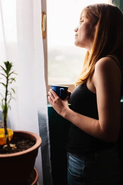 Jeune femme avec une tasse debout près de la fenêtre profitant de détente dans sa maison — Photo