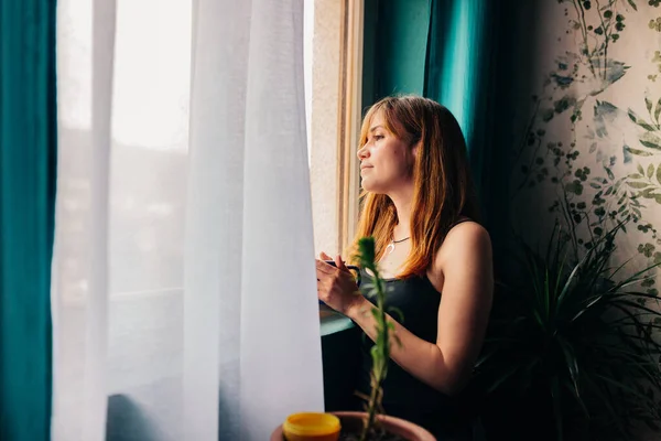 Молода жінка з чашкою, що стоїть біля вікна, насолоджується відпочинком у її будинку — стокове фото