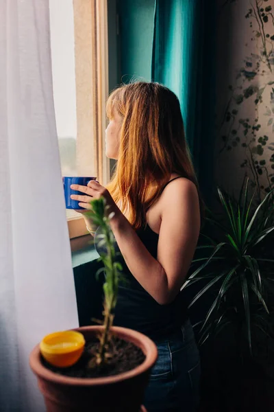 Ung kvinna med en kopp stående nära fönstret njuter av avkoppling i sitt hem — Stockfoto