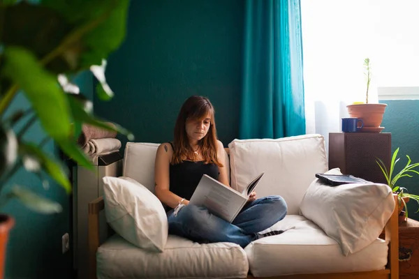 Young red-haired woman, reading a book in a couch surrounded by plants. Leisure indoors entertainment.