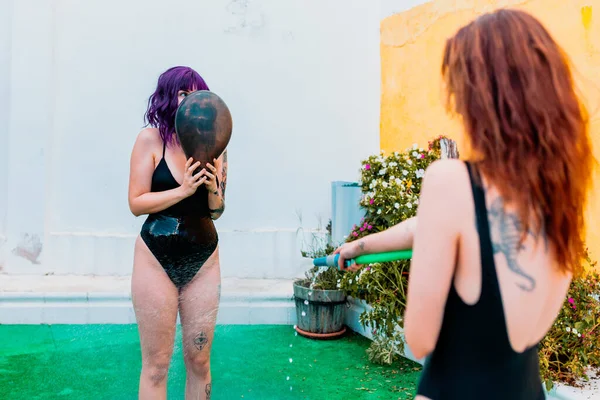 Dos mujeres felices riendo en traje de baño jugando bajo gotitas de agua de una manguera en el patio trasero —  Fotos de Stock