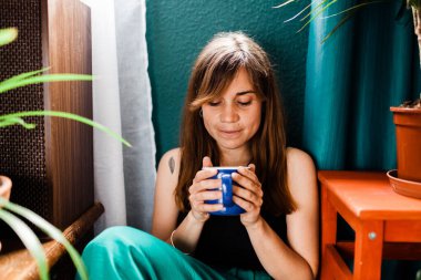 Kızıl saçlı kadın evde oturup yerde kahve içiyor..