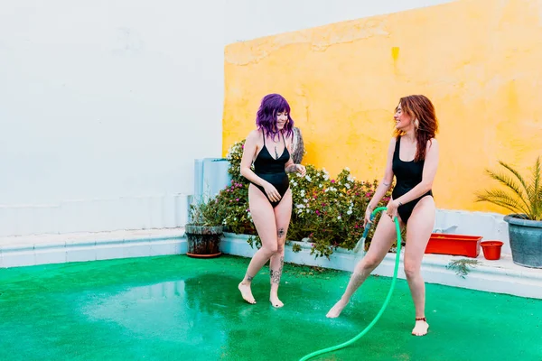 Dos mujeres felices riendo en traje de baño jugando bajo gotitas de agua de una manguera en el patio trasero —  Fotos de Stock