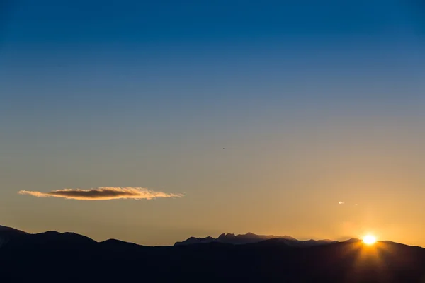 Soleil derrière des silhouettes sombres de montagne, avec un ciel bleu et orange — Photo
