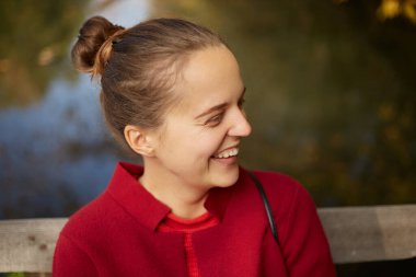 Duygusal olarak gülen genç bir bayanın nehir kıyısında açık havada oturması, kırmızı gömlek giyen mutlu bir bayanın yan görünüşü, olumlu duygular ifade ediyor..