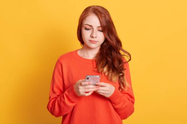 Ciddi kızıl saçlı, turuncu kazaklı bir kadın, sarı duvarda izole edilmiş poz veriyor, cep telefonu kullanan düşünceli bir kız, SMS yazıyor ya da sosyal ağları kontrol ediyor..