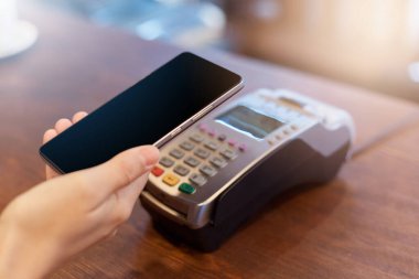Yüzsüz müşteri NFC Teknolojisi ile ödeme yapıyor, cep telefonu ile temas kurmadan ödeme yapıyor, dükkanda ya da kafede ahşap tezgahın üzerinde terminal.