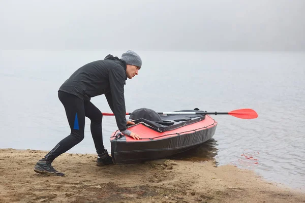 Lonely man going paddling on kayak in early foggy morning, sports man being fond of water sport, guy pushing boat into water, looks at misty river, wearing warm clothing.