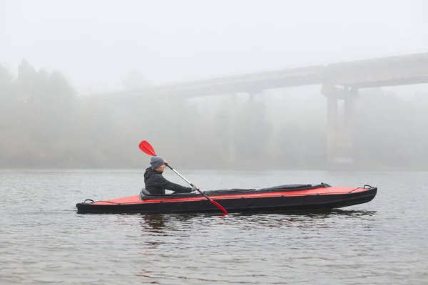 川での若いカヤックのサイドビュー パディング 背景に橋で霧の朝にウォータースポーツを楽しむ男 寒い秋の日に男性ボートを漕ぐ — ストック写真