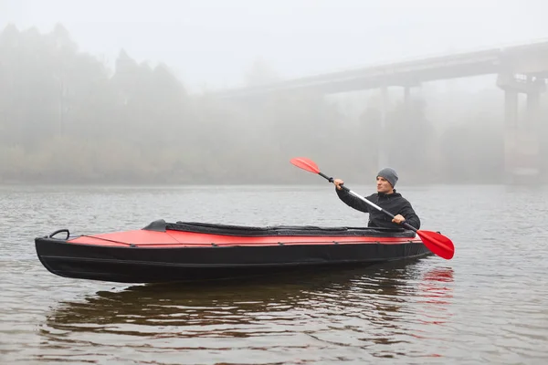 英俊的小伙子 手里拿着桨 划着船 遥望远方 秋天的早晨 雾蒙蒙的 皮划艇在河中央 背景是一座桥 — 图库照片