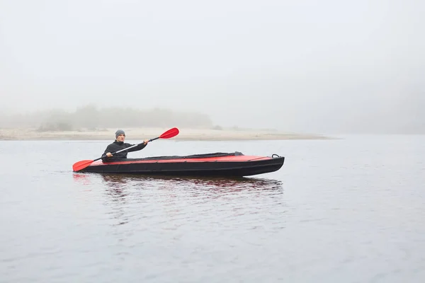 年轻迷人的小伙子穿着温暖的黑色夹克 在雾蒙蒙的早晨划艇 遥望远方 享受着他的业余爱好 喜欢水上运动 坐在河中央的小船上 — 图库照片