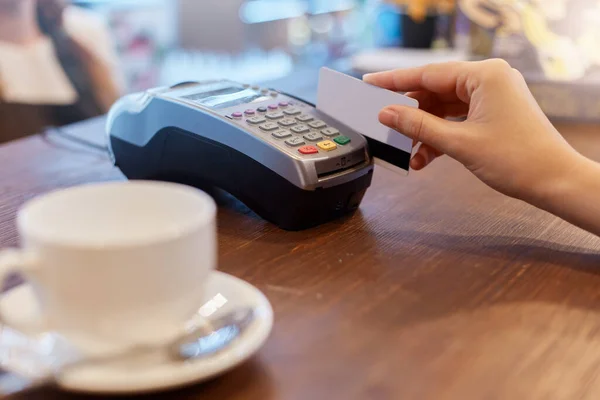 Paying with credit card for purchase in cafeteria, faceless woman using bank card for pay, wooden counter in cafe with terminal and white ceramic cup with coffee or tea.