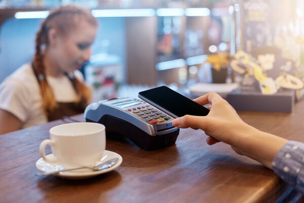 Faceless woman customer paying in cafeteria via contactless way, unknown female holding smart hone near terminal with waiter on background, white cap with coffee on wooden counter.