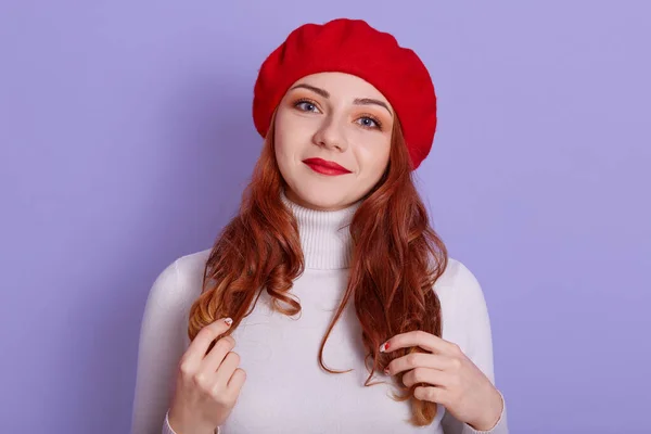 Positive adorable female model smiles, keeps hands on her curls, wears old fashionable beret and white sweater, lady with perfect skin and red lips looks at camera.