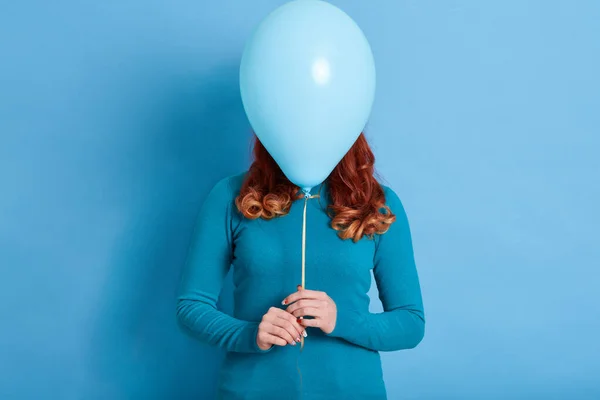 Portrait of woman hides her face behind helium balloon, red haired female with curls holding ballon in front her face against blue background, holiday, celebration.