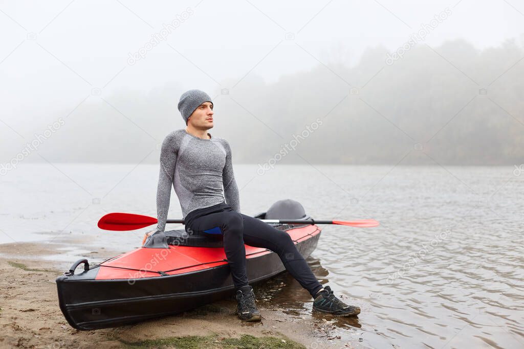 Adult handsome man sitting on boat and looks into distance with dreamily facial expression, wearing sporty wear,posing with foggy river on background.