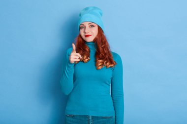 Neşeli güzel Avrupalı bayan kameraya silah hareketi yapıyor, tercihini ifade ediyor, geniş bir şekilde gülümsüyor, kırmızı dudaklar giyiyor, kazak ve şapka giymiş, mavi arka planda soyutlanmış..