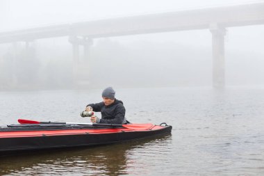 Nehrin ortasında kanoyla gezen yakışıklı, termostan sıcak bir içecek dolduran, siyah ceketli, gri şapkalı, güzel, sisli sonbahar gününde kanoda çay ya da kahvenin keyfini çıkaran genç bir erkek..