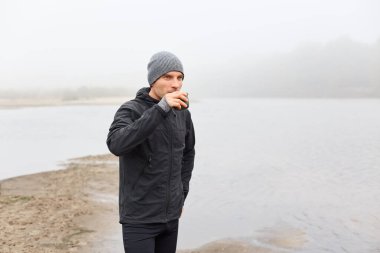 Beyaz adam sonbaharda nehrin kıyısında durur ve çay ya da kahve içer. Arkaplanda mesafeye, nehre ve bulutlu gökyüzüne bakan yakışıklı adam..