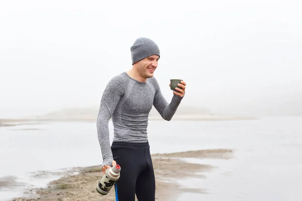 Handsome man drinking hot coffee from thermos, smiling, laughing. Beautiful view of foggy river in cloudy autumn morning, happy guy wearing gray shirt and black trousers.