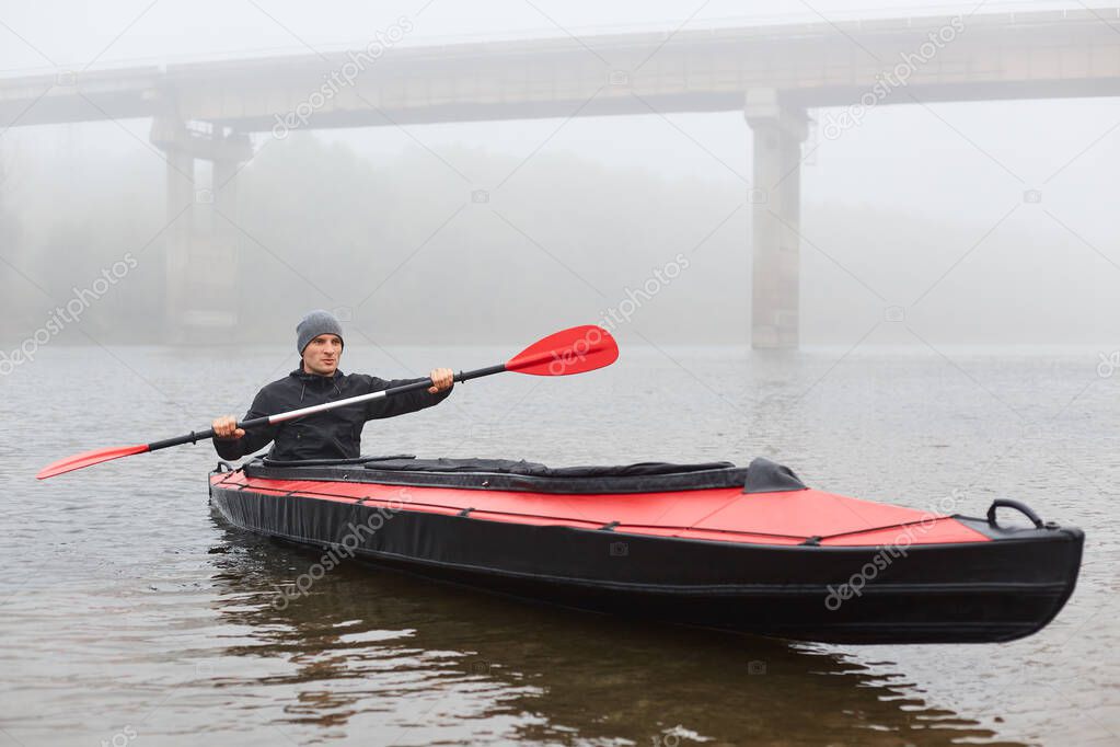 Front view of man paddles in kayaks on calm lake or river near bridge in cloudy autumn day, guy looks straight ahead, holding oar in hands and rowing, wearing black jacket and cap.
