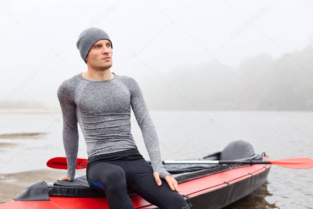 Portrait of man grey cap and shirt posing on bank on river in boat, looking dreamily in distance, being serious and concentrated, having rest after rowing boat.