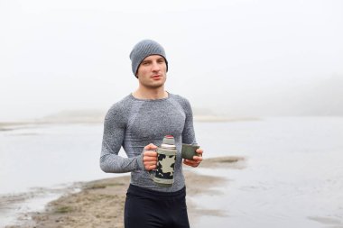 Nehir kıyısında yakışıklı, sisli gölün yanında termosla poz veren yakışıklı bir adam, sıcak kahveyi ve çayı seven, şapkalı, gri gömlekli ve pantolonlu, kameraya bakan dalgın bir adam..