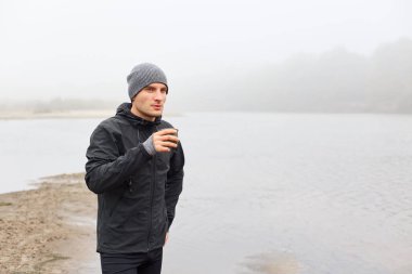 Yakışıklı, düşünceli bir adam elinde kahve ya da çayla gölün kenarında dikiliyor, dalgın bakışlarıyla, siyah ceketli ve gri şapkalı, nehir kıyısında duruyor..