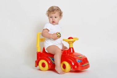 Küçük Avrupalı çocuk çıplak ayaklı, beyaz takım elbiseli, kıvırcık saçlı sarışın çocuk başka tarafa bakıyor, ilginç bir şey görüyor, oyuncak arabayı elinde tutuyor..