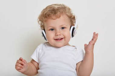 Beyaz arka planda, kulaklıklı, kameraya mutlu bir gülümsemeyle bakan, gizli dişlerini gösteren, tişört giyen, sarışın, kıvırcık saçlı ve elini kaldıran bir çocuk şarkı dinliyor..