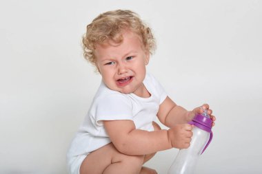 Beyaz arka planda çömelmiş ağlayan bir çocuk, içmek isteyen bir çocuk, elinde boş bir bardak, güzel sarı dalgalı saçlı bir bebek, susamış hissediyor..